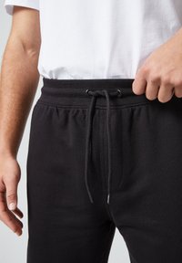 Zwarte joggingbroek met een trekkoord in de taille, met een zachte textuur en zijzakken. Gedeeltelijk gedragen met een eenvoudig wit T-shirt.