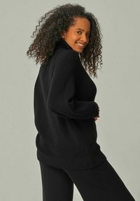 Schwarzer Strickpullover mit hohem Kragen und lockeren Ärmeln, ausgestattet mit geripptem Textur und lässiger Passform, kombiniert mit schwarzen Hosen.