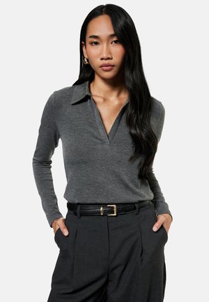 Donna con lunghi capelli neri che indossa una camicia grigia a maniche lunghe con colletto, pantaloni neri e orecchini a cerchio dorati, con le mani nelle tasche.