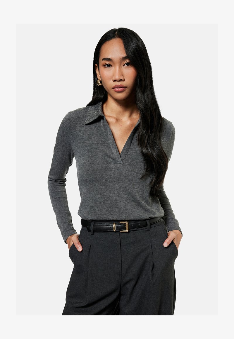 Mujer con cabello largo y negro, que lleva una camisa gris de manga larga con cuello, pantalones negros y pendientes de aro dorados, con las manos en los bolsillos.