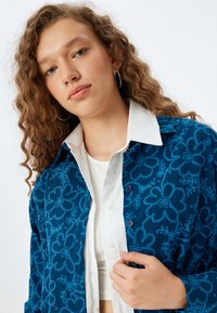 Långärmad skjorta som har ett marinblått tyg med ljusblå blommönster, en vit krage och knappar längs fram.