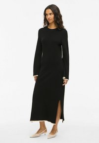 Černé šaty s dlouhými rukávy se střihem do přímého střihu, boční rozparkou a krémovým lemem na manžetách i spodním okraji. Materiál vypadá jako pletený s hladkou texturou.