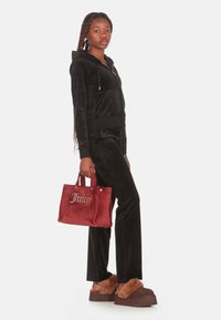 Survêtement en velours noir avec un hoodie zippé, associé à un sac à main en velours rouge portant le logo "Juicy", et des sandales à semelle fourrée marron.