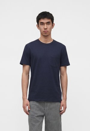Młody mężczyzna stojący przodem, ubrany w granatowy t-shirt z kieszeniami oraz szare spodnie w paski na jednolitym jasnym tle.