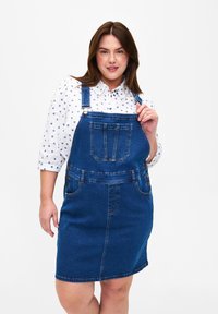 Denim haljina na bretele u tamnoplavoj boji s podesivim trakama, prednjim džepom i detaljima gumba sa strane. Nosena preko bijele košulje s malim plavim uzorcima srca.