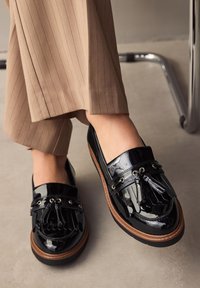 Mocassins en cuir verni noir au design structuré, ornés de pampilles décoratives et d'accents métalliques, reposant sur une semelle marron contrastante.