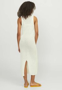 Vestido sin mangas de color crema, con longitud hasta la rodilla y un patrón de tejido texturizado, con una abertura en la parte trasera central, combinado con chanclas amarillas.