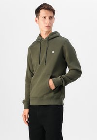 Felpa verde oliva in morbido tessuto, con tasca a marsupio, cappuccio regolabile con cordino e polsini a costine. Pantaloni neri.