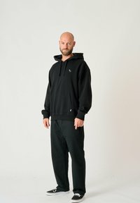 Black hooded sweatshirt with a front pocket and drawstrings, featuring a small gray bird logo. Worn with loose black pants and black shoes.