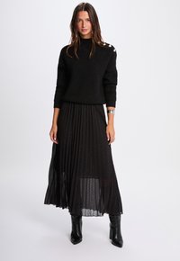 Sort striktrøje med høj krave og knapdetaljer. Plisseret sort nederdel med gennemsigtigt stof og subtile glitrende elementer. Knæhøje sorte støvler.