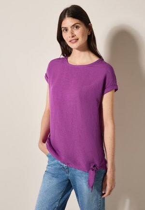 Chemise violette à manches courtes avec un col rond et un détail de nœud en bas à gauche. Fabriquée en tissu léger et doux.