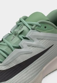 Zbliżenie na jasnozielony i szary but do biegania marki Nike z czarnym logo Swoosh i napisem „Vapor Pro” na języku.