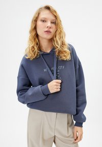 Felpa blu navy con una stampa frontale che riporta la scritta "New York City." Presenta un cappuccio con cordino e polsini a costine, abbinata a pantaloni beige.