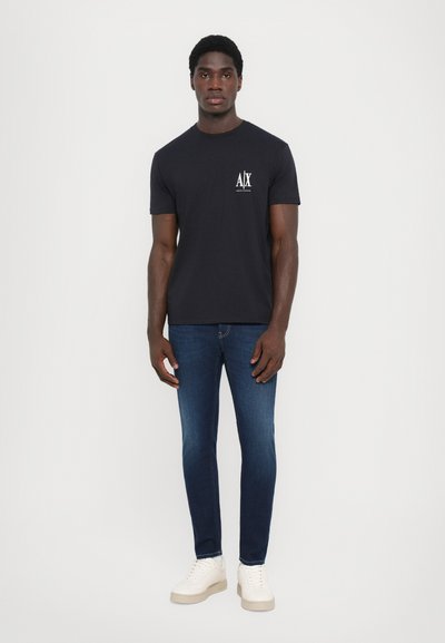 Tricou din bumbac de culoare bleumarin, cu mâneci scurte și un logo pe față. Asortat cu blugi închis la culoare, cu croi slim, și adidași albi. Fundal simplu.