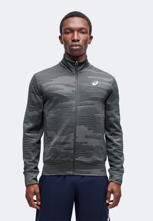 Mann trägt eine graue, gemusterte Sportjacke mit Reißverschluss und hohem Kragen sowie dunkelblaue Sportshorts und steht vor einem einfarbigen Hintergrund.