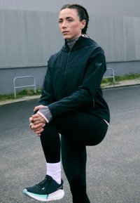 Črna športna jakna z zadrgo, sivo dolgo majico spodaj, črne legice in sivo-črne tekaške čevlje z belimi podplati, oblečene z belimi nogavicami.