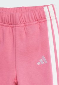 Ružové tepláky s elastickým pásom, so bielymi vertikálnymi pruhmi po stranách a strieborným logom na spodnej časti nohy.