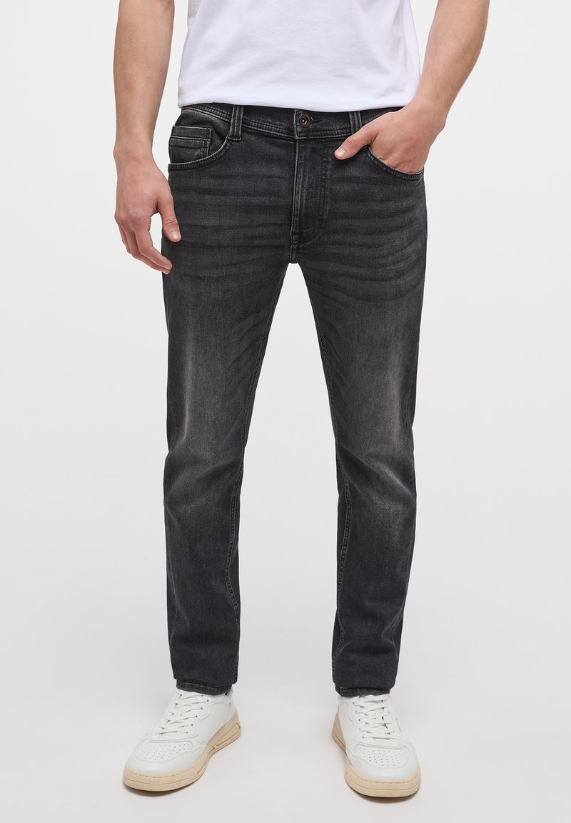 Homme portant un jean noir slim, des baskets blanches et un t-shirt blanc, main dans la poche, debout devant un fond clair uni.