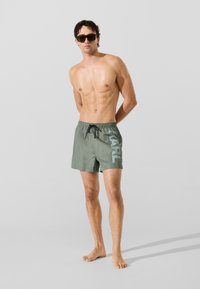 Póló nélküli férfi sötétzöld fürdőnadrágot visel, amelyen zsinór van, és az oldalukon nagy felirattal "KARL" áll, mezítláb egy sima szürke háttér előtt.