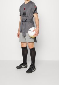 Grå Nike sporttröja med svarta accenter, grå shorts, svarta knähöga strumpor och svarta fotbollsskor som håller en vit fotboll.