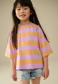 T-shirt oversized à manches courtes avec des rayures horizontales violettes et oranges, ornée de broderies florales colorées. Assorti avec un short en denim déchiré.