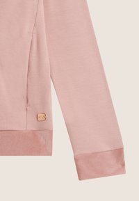 Tissu de sweat-shirt rose clair avec une texture douce, présentant un ourlet et des poignets côtelés. Comprend un petit détail de logo en relief.