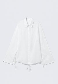 Hvid bomulds skjorte med krave, lange ærmer og justerbare bind ved håndleddene. Knaplukning fortil og en let løs pasform.