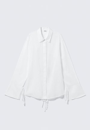 Witte katoenen shirt met een kraag, lange mouwen, en verstelbare strikjes bij de manchetten. Knoping aan de voorkant en een iets losse pasvorm.