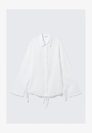 Chemise en coton blanche avec un col, des manches longues et des liens réglables aux poignets. Fermeture par boutons sur le devant et coupe légèrement ample.