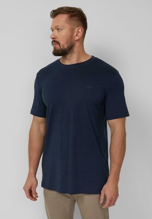 Mann mit Bart, der ein schlichtes marineblaues kurzärmeliges T-Shirt und beige Hosen trägt und auf einem hellgrauen Hintergrund nach links schaut.