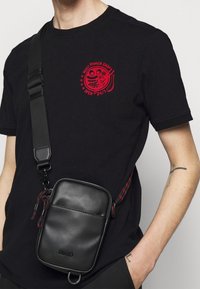 Sac bandoulière en cuir noir avec une sangle à motif rouge et noir, doté d'une fermeture éclair, d'une poche avant et d'un logo en relief à l'avant.