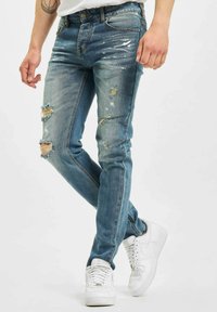Blå denimjeans med slitte detaljer, falming og rifte aksenter, kombinert med hvite joggesko. Slim fit design, mid-rise liv.