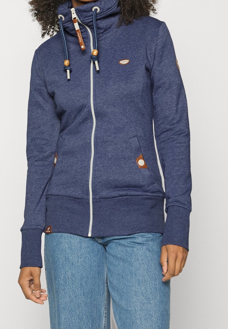 Sweat à capuche zippé bleu marine en tissu doux, doté d'un col montant, de cordons contrastants et de deux poches latérales avec des accents marron.