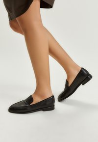 Mocassini in pelle nera con tacco basso, caratterizzati da un design con ritagli e una texture liscia. Le scarpe sono indossate con caviglie scoperte.