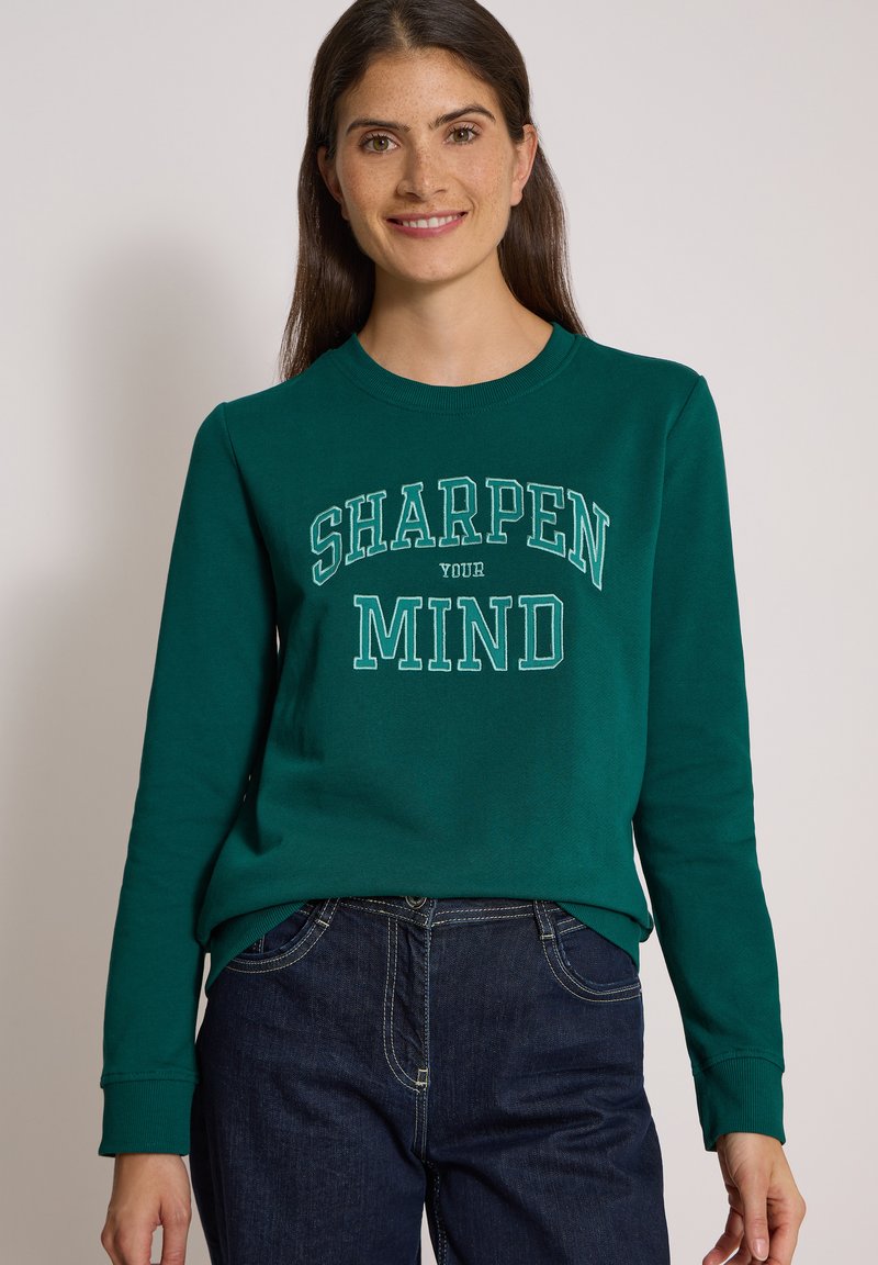 Femme aux longs cheveux foncés portant un sweat vert avec le texte "SHARPEN YOUR MIND" et un jean bleu foncé, souriante à la caméra.