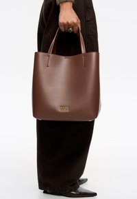 Bolso tote de piel marrón con una textura suave, que cuenta con dos asas para el hombro y un detalle del logo dorado en la parte inferior. Diseño sencillo y abierto.