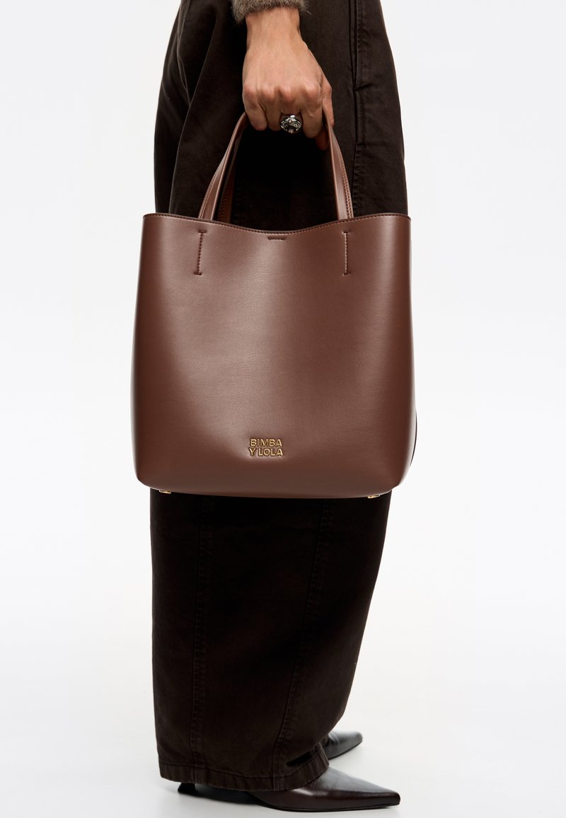 Bolso tote de piel marrón con una textura suave, que cuenta con dos asas para el hombro y un detalle del logo dorado en la parte inferior. Diseño sencillo y abierto.
