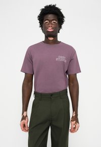 Camiseta morada de manga corta y corte relajado con el logo "OBEY STUDIOS" impreso en el lado izquierdo del pecho, combinada con pantalones oscuros verdes.