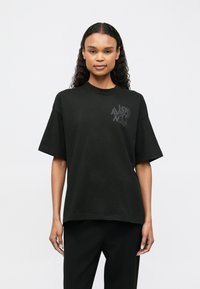 Melnas kokvilnas T-krekls ar apaļu kaklu un pārmērīgu piegriezumu. Ar smalku grafisku logotipu kreisajā krūtī. Īsas piedurknes un gluda tekstūra.