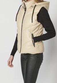 Veste matelassée beige sans manches à capuche avec fermeture éclair devant et poches zippées, portée sur un top noir à manches longues et un pantalon noir.