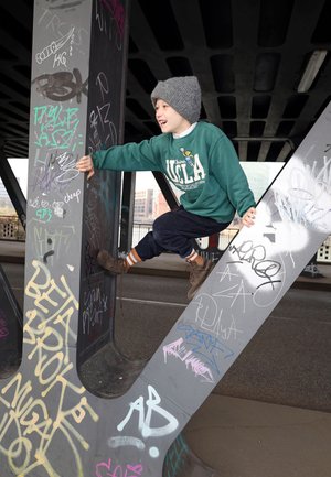 Kind in groene UCLA-trui en grijze beanie beklimt stalen balken bedekt met graffiti onder een brug, glimlachend en naar rechts kijkend.