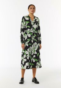 Blumenkleid mit schwarzem Grund, das große weiße und grüne Blumen zeigt. Lange Ärmel, V-Ausschnitt und eine lockere, mittellange Silhouette. Flache Schuhe.