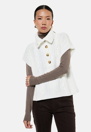 Młoda kobieta ubrana w biały krótki sweter warkoczowy z dużymi guzikami, na brązowym golfie z długim rękawem i ciemnych spodniach.