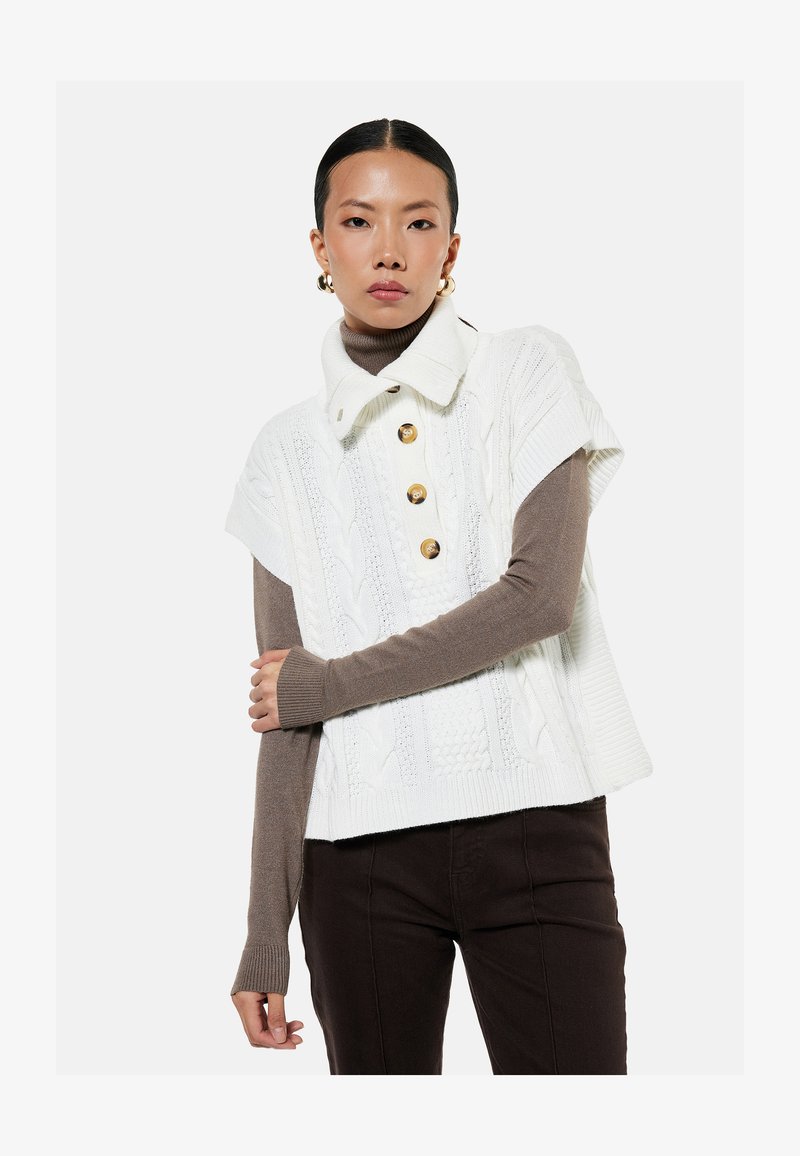 Młoda kobieta ubrana w biały krótki sweter warkoczowy z dużymi guzikami, na brązowym golfie z długim rękawem i ciemnych spodniach.