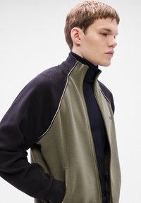 Olívazöld testű és fekete ujjú szabadidődzseki, magas nyakkal és fehér szegéllyel a cipzár mentén, puha anyagból készült.