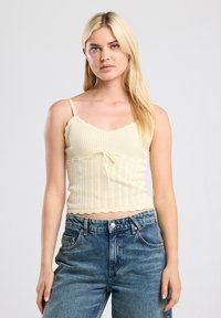 Yellow knitted crop top with a scalloped neckline and hem, featuring adjustable straps and a central bow detail. Paired with blue denim jeans.