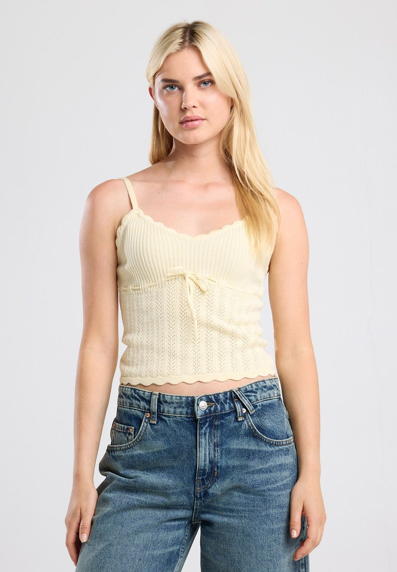 Yellow knitted crop top with a scalloped neckline and hem, featuring adjustable straps and a central bow detail. Paired with blue denim jeans.