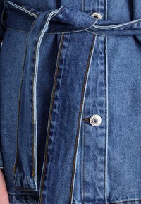 Denim jakk detail, millel on lipsu vöö, hõbedased kinnitusdetailid ja kontrastsete õmblustega tekstureeritud keskmise sinise kangaga.