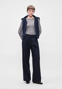 Top de manga longa cinza, colete sem mangas azul-marinho e calças largas azul-marinho com riscas. Tecido texturizado com detalhes em botão. Sapatos pretos.