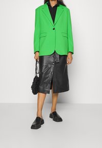 Blazer sovradimensionato verde brillante, gonna in pelle nera con bottoni, scarpe nere slip-on e una borsa nera texturizzata. Linee pulite, look moderno.